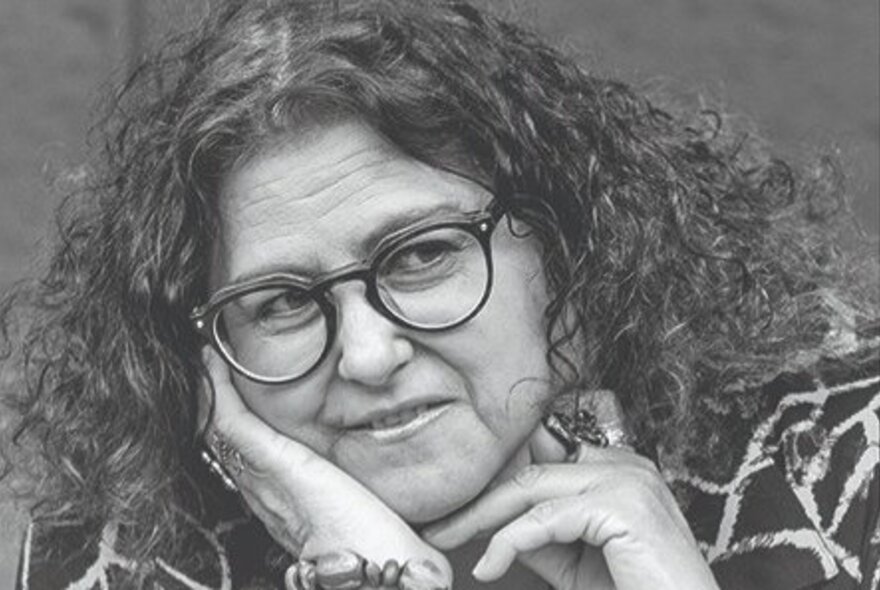 A woman, resting her cheek on her hand, her curly hair framing her face, wearing glasses and a chunky bracelet around her wrist.