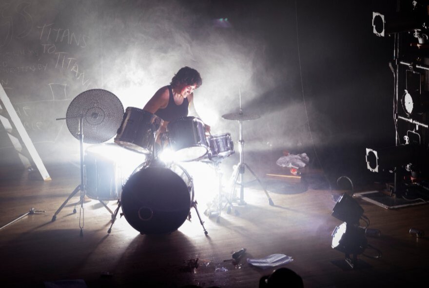 An artist performing a drum set solo on a dark, smoky stage, heavily backlit by bright stage lights creating a silhouette and dramatic flare.