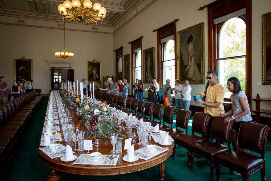 Very long State Apartment dining table set for a formal dinner with visitors looking on, taking photos, in a formal room with chandelier and portraits.