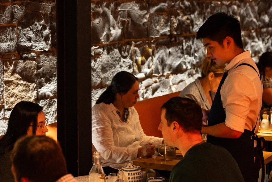 Diners at tables in a restaurant and a waiter standing listening. 