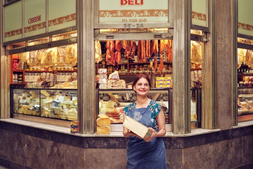 Smiling tour guide wearing a blue apron, holding a large piece of hard cheese and standing in front of a deli stall at Queen Victoria Market.