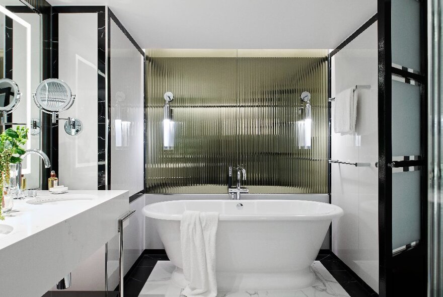 A bathroom at the Royce Hotel with a white freestanding bathtub, and white, glass, chrome and timber fixtures.