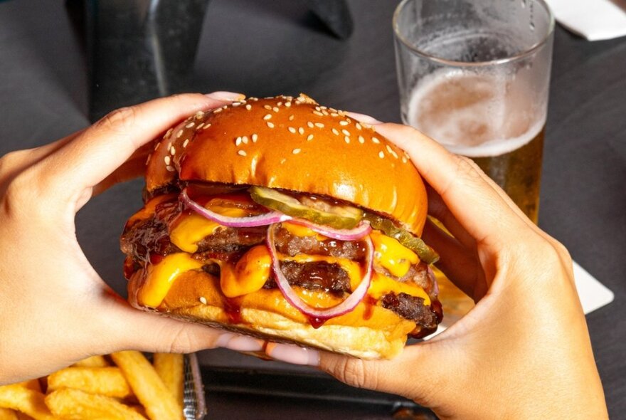 Hands holding a large hamburger with onion and cheese next to chips and half a glass of beer.