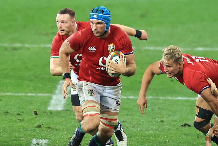 Lions rugby players wearing red tops and white shorts playing rugby on a green sports field, one of them carrying the ball.