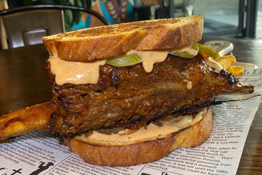 A toasted sandwich with a giant single beef rib, mayonnaise and pickles, resting on a printed sheet of paper. 