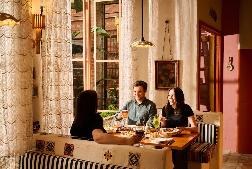 Interior of restaurant with three diners seated at a booth table eating and drinking.