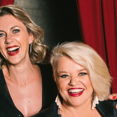 Singers Amelia Ryan and Libby O’Donovan, both wearing dark tops and smiling, posed against a red velvet curtain.
