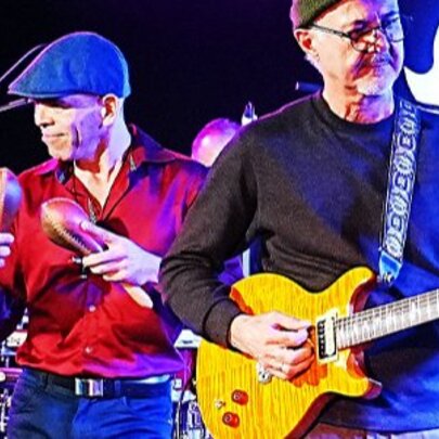 Two musicians in a band performing live on stage, one playing a guitar and the other shaking maracas.