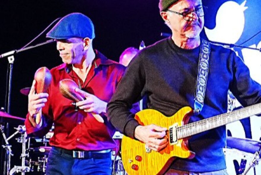 Two musicians in a band performing live on stage, one playing a guitar and the other shaking maracas.