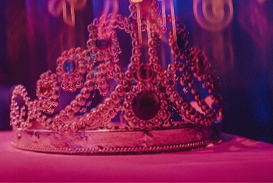 An elaborate silver tiara with gems resting on a pink surface, lit up by a pink neon light.