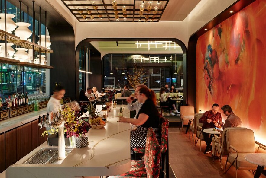 Long view of restaurant interior, with patrons seated at high stools at the marble bar, the bartender, and two people seated at a small table against a wall painted with a mural.