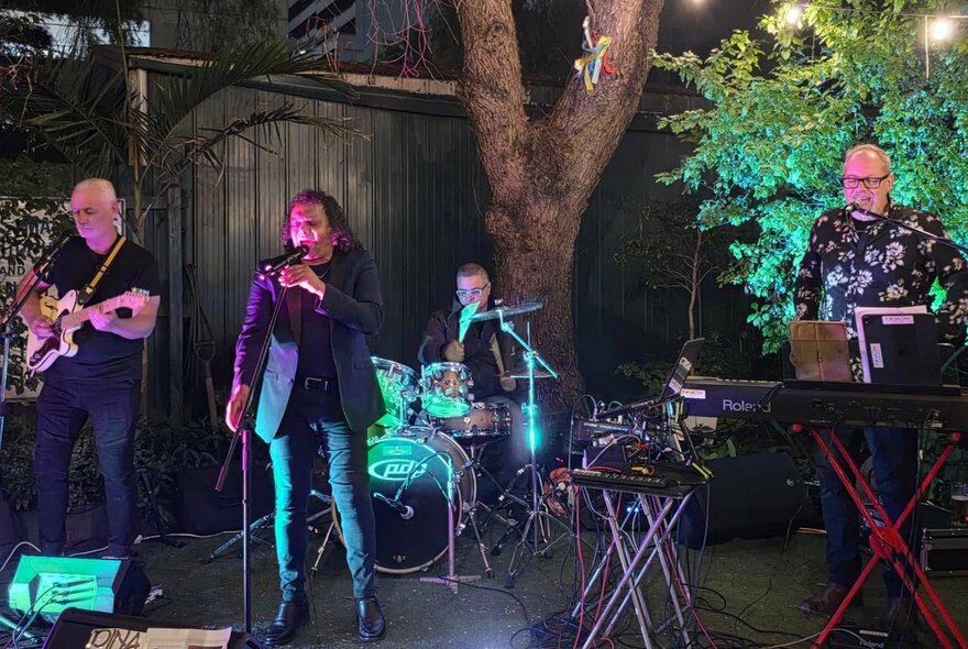 Soul musician Dinash Daniel playing with a band in a small garden area with a large tree trunk in the background. 
