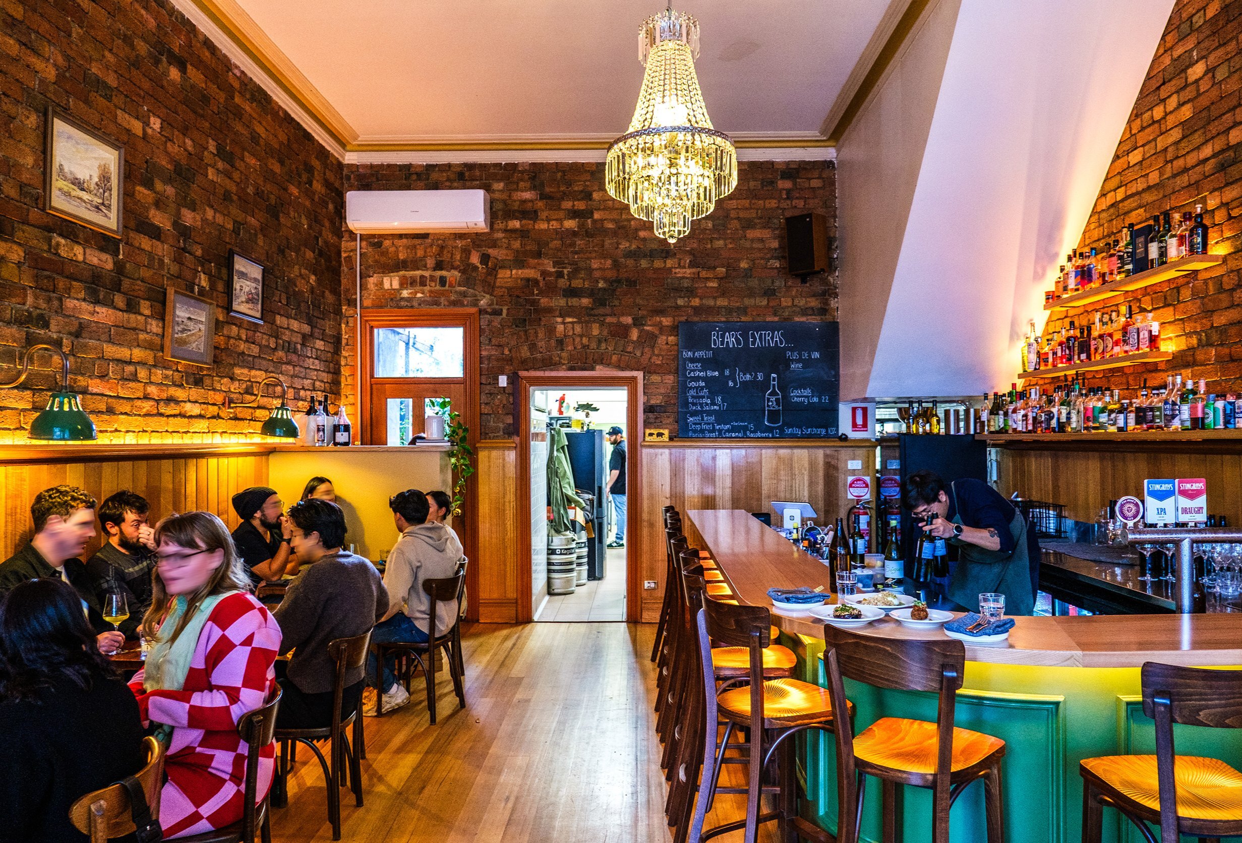 A group of customers area seated at a cosy wine bar with brick walls and a chandelier hanging from the ceiling. There is a bar tender behind the bar.