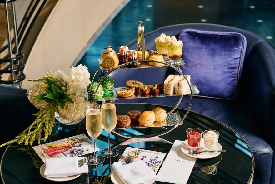A round table set for a high teas service with flowers, savoury and sweet food, and two glasses of champagne.