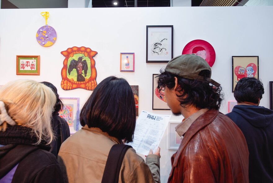 An exhibition of small wall-based artworks in a gallery space, with people in the foreground viewing the works.
