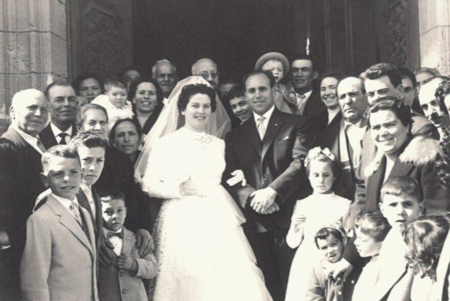 A black and white wedding photo with many family members crowded around the bride and groom outside a church. 
