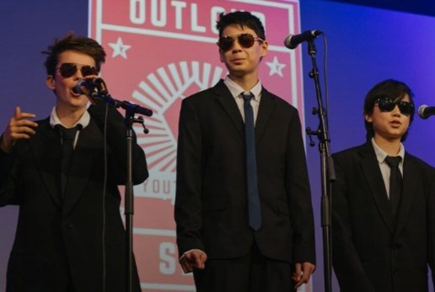 Three teenage boys on a stage behind microphone stands, wearing dark suits, ties, and dark glasses as they recite original poetry.