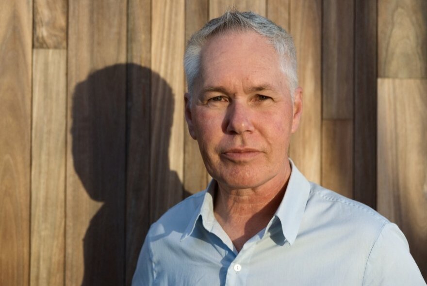 Older man with grey hair, wearing a pale blue shirt, standing next to a wooden fence.