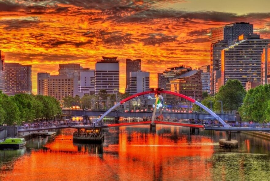 A vibrantly hued sunset, reflected on the surface of Melbourne's Yarra River with a footbridge in the foreground and Melbourne's skyline in the background.