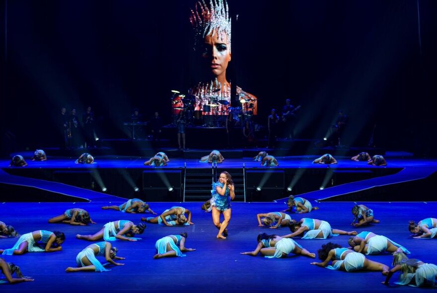 Dancers prostrate on the ground in front of a single large figure on a screen.