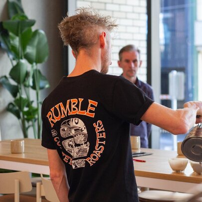 A person hosting a coffee cupping demonstration in a shop, pouring coffee into cups lined up on a counter, with participants watching.