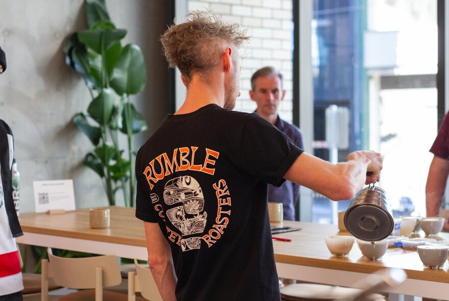 A person hosting a coffee cupping demonstration in a shop, pouring coffee into cups lined up on a counter, with participants watching.