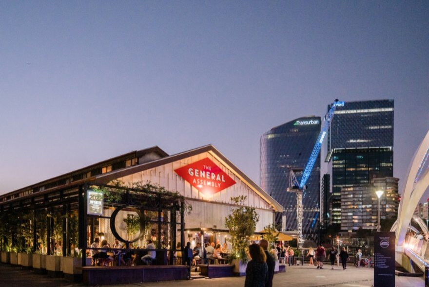 The exterior of General Assembly bar at twilight, with a fading sky and two city skyscrapers in the background.