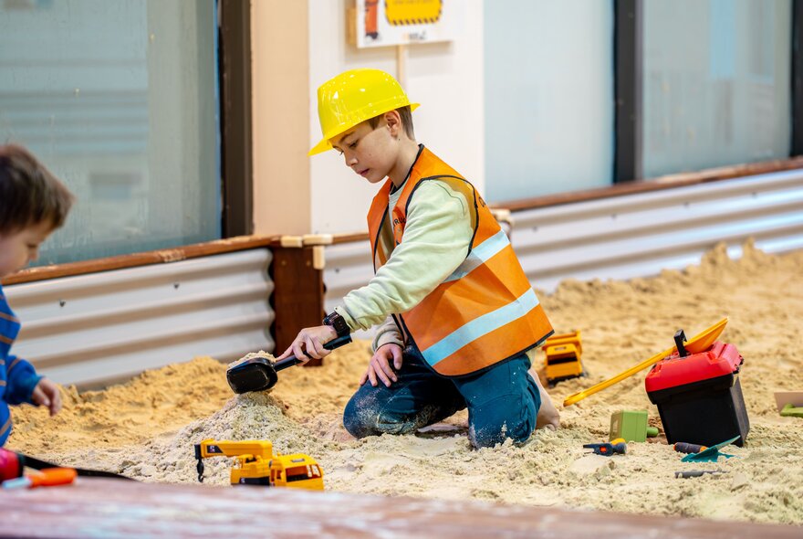 A child playing in a large indoor sandpit filled with diggers and shovels.