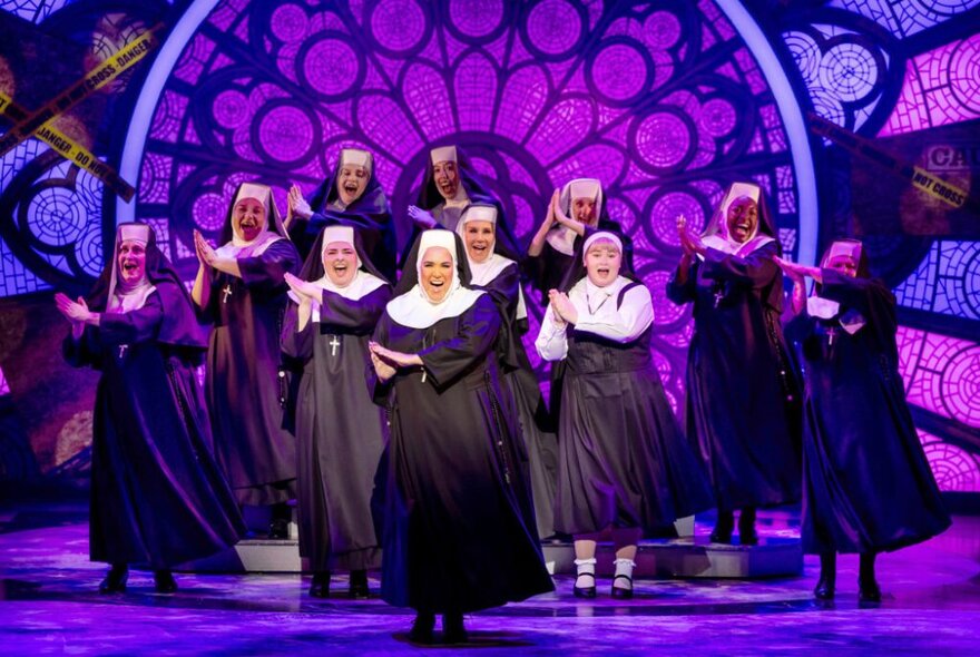 A group of nuns in black and white habits, singing on stage with a large purple stained glass set decoration behind them.