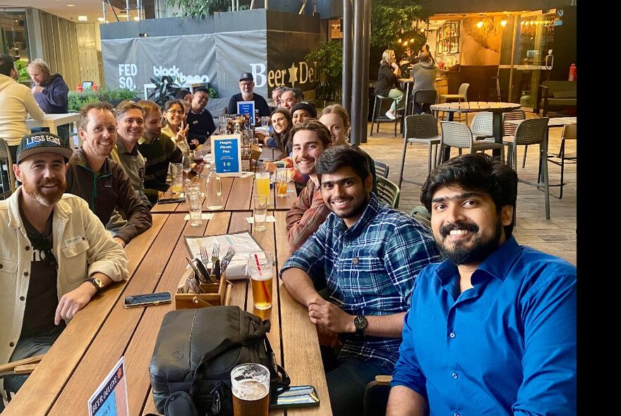 A group of people sitting around a table at an outdoor courtyard in a pub, all looking smiling and looking at the viewer.