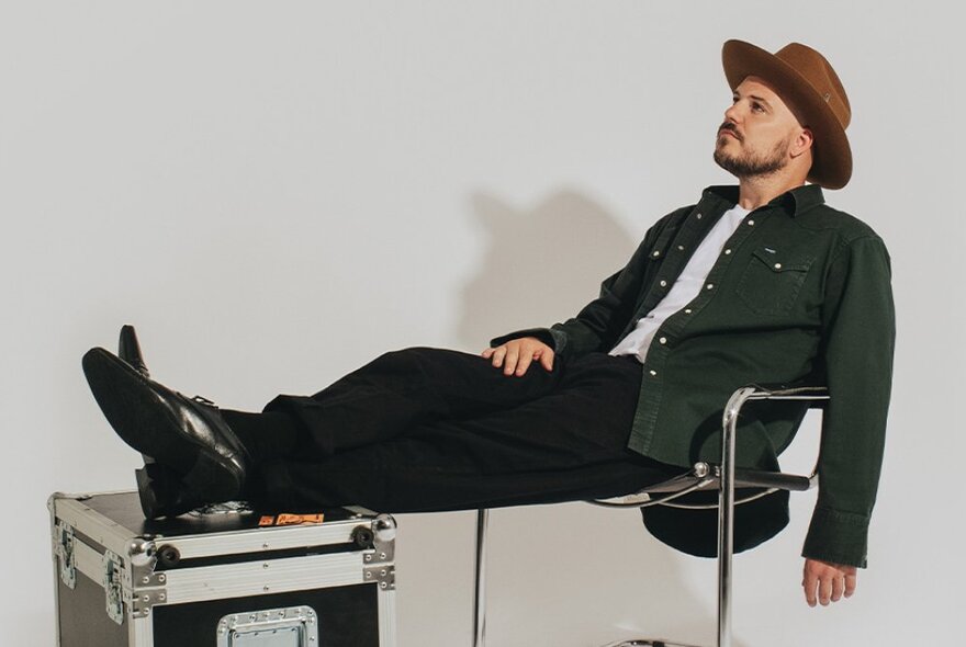 Bearded country singer Taylor Sheridan, seated with his feet on a music case, wearing a hat and denim shirt.