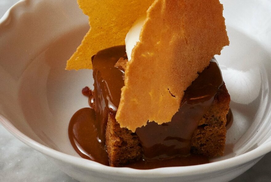 A serve of pudding in a white bowl, with a sauce on top of it, plus wafers and ice-cream. 