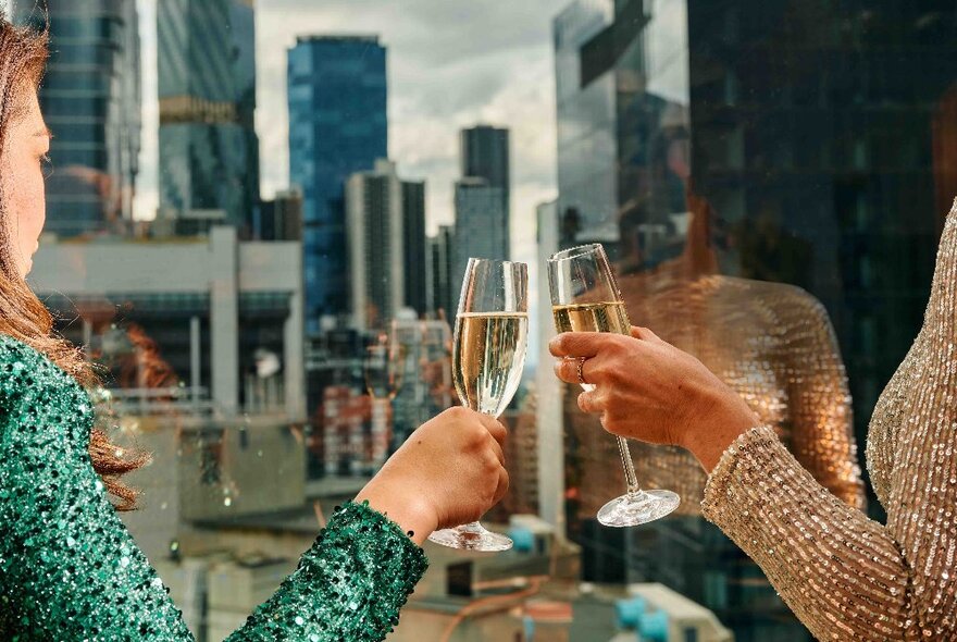 Two people clinking glasses with two filled champagne flutes, with views of city buildings through the large window behind them.