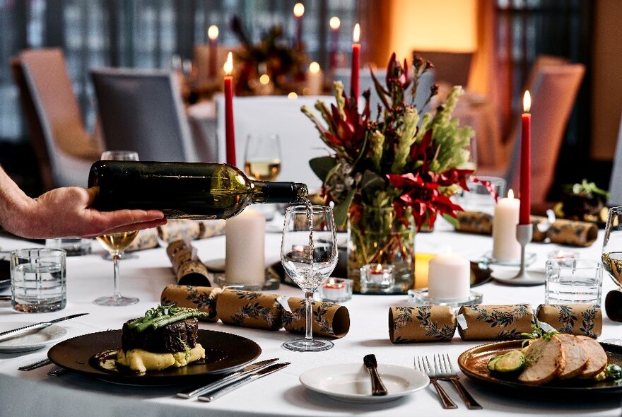 A table in a restaurant with plates of food, wine being poured into a glass, candles and festive table decorations.