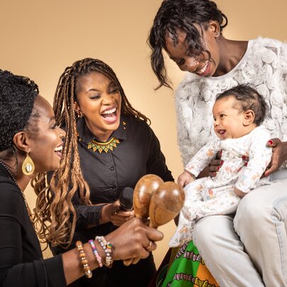 Three people interacting with a baby that is seated on a women's lap.