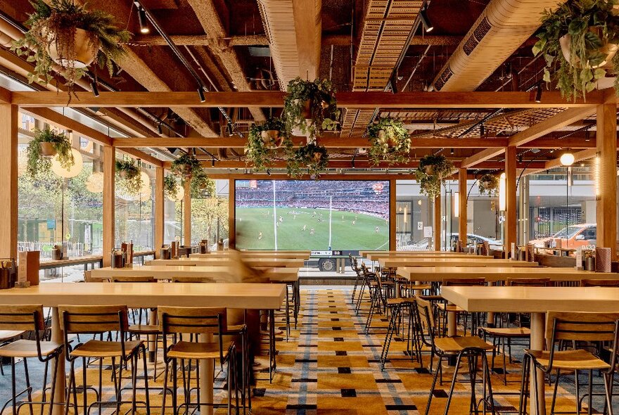 Interior of the Quarterhouse, with exposed timber ceiling beams, hanging baskets of plants, rows of high tables with stools, all facing a large LED screen on the rear wall.
