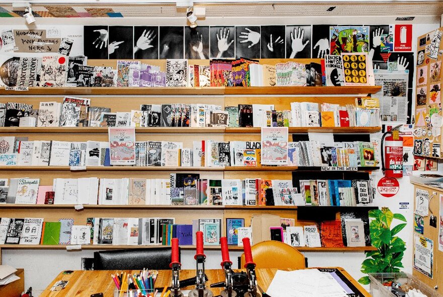 The studio space at Sticky Institute, with a wall of shelves against the rear wall, and a large work bench with paper, and tools for making zines.