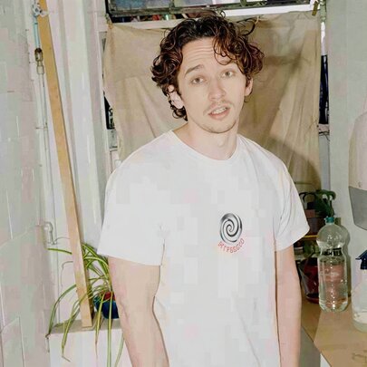 Comedian Milo Edwards wearing a white t-shirt, standing inside a bathroom, looking directly at the camera with a neutral expression.