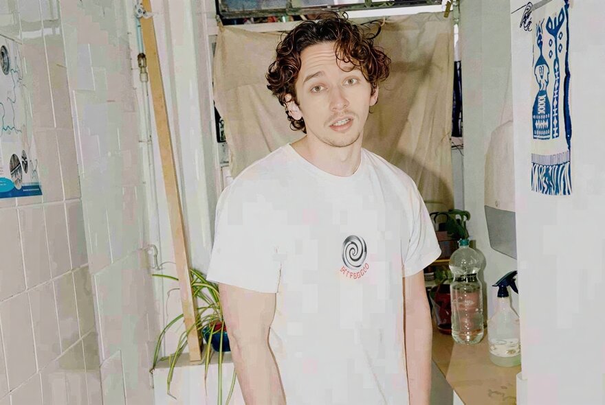 Comedian Milo Edwards wearing a white t-shirt, standing inside a bathroom, looking directly at the camera with a neutral expression.