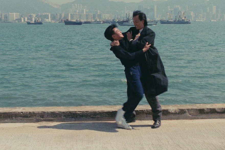 A scene from the 1986 movie A Better Tomorrow, featuring two characters fighting each other near the waterfront, boats and a city skyline on the horizon.