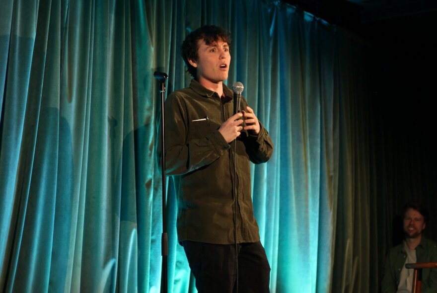 A stand-up comedian talking into a microphone they hold in their hand, performing live on stage.