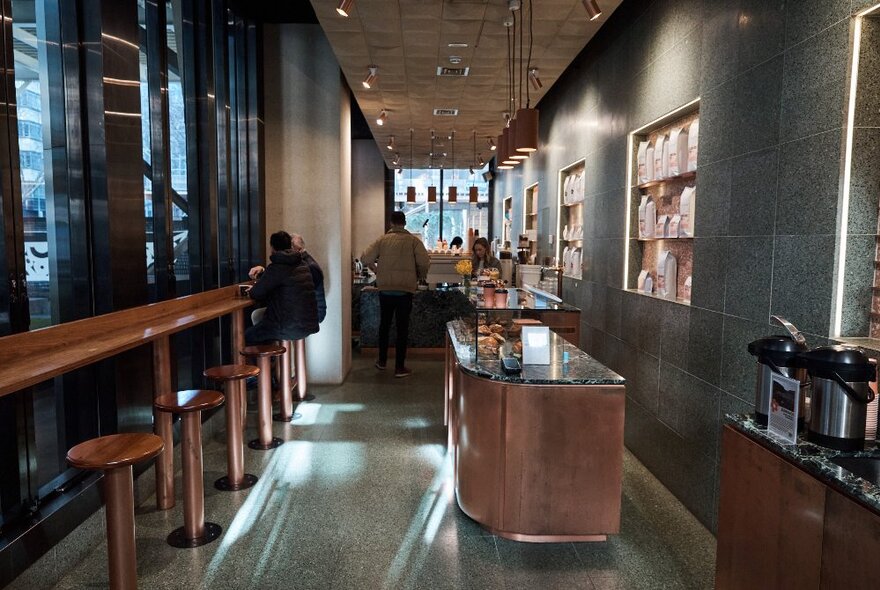 The internal view of a green-tiled cafe with a long timber bench and stools, an island bench and a counter at the back.