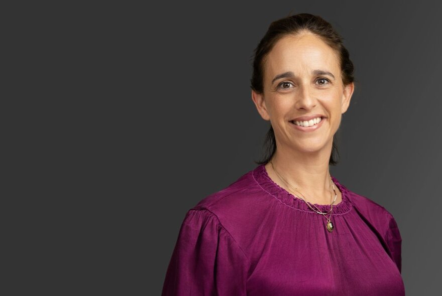 Smiling head and shoulders of Daniela Gavshon, a woman wearing a dark purple blouse posed against a dark background.