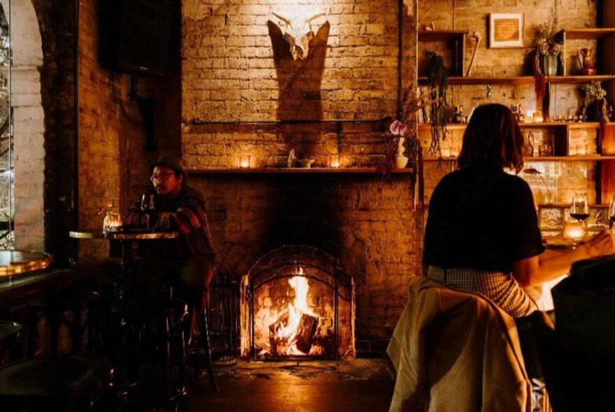 People sitting beside the fire in a cosy bar with brick walls and vintage trinkets on the shelves.