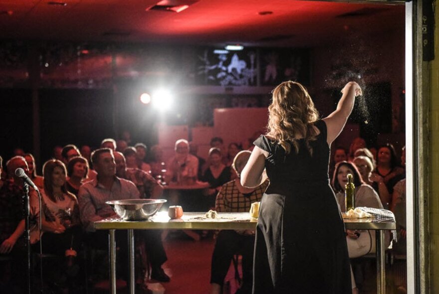 A woman performing a cooking demonstration in front of a small audience in an intimate setting. 