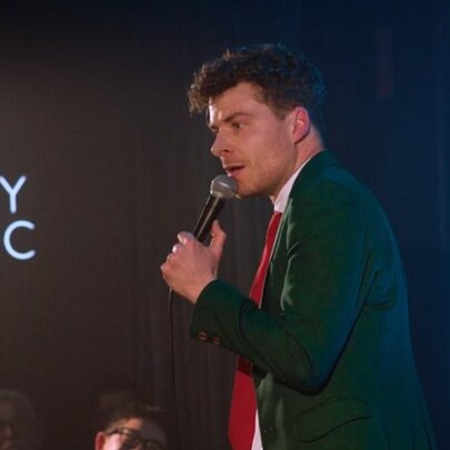 Comedian Oliver Coleman wearing a suit jacket and red tie, holding a microphone and performing stand-up comedy on a stage at Comedy Republic.