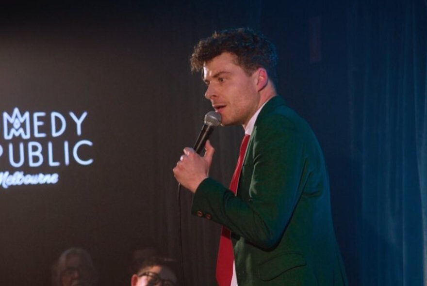 Comedian Oliver Coleman wearing a suit jacket and red tie, holding a microphone and performing stand-up comedy on a stage at Comedy Republic.
