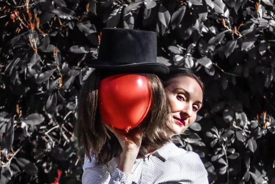 A person playfully hiding their face behind a red balloon topped with a black felt hat.
