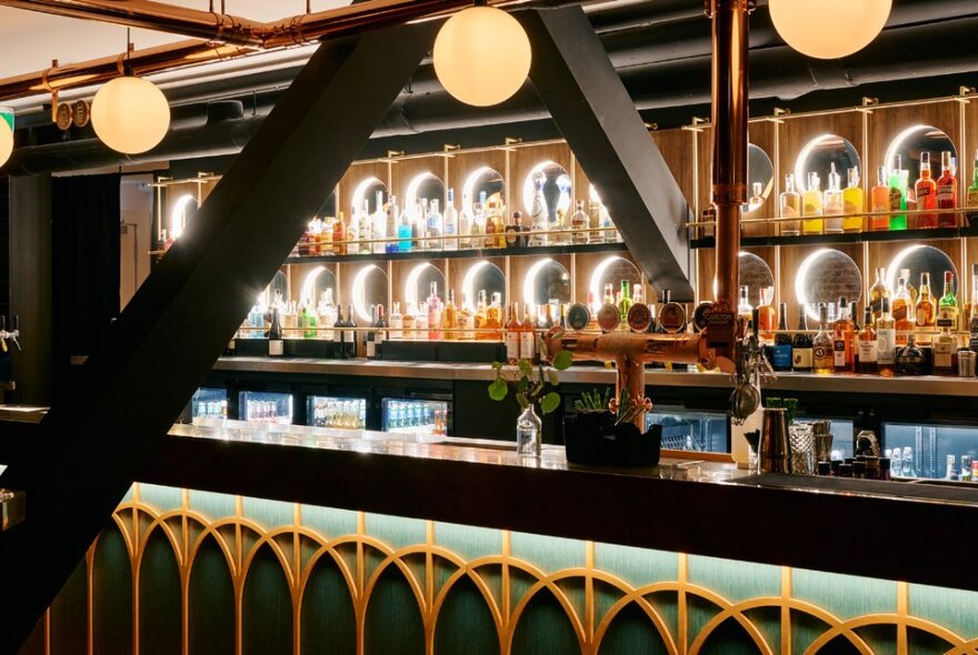 Cocktail bar counter with taps, fridges and glass shelves lined with bottles.