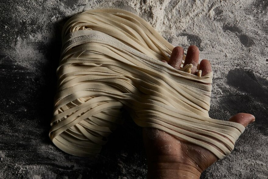 Hand made noodles on a floured worktop, a hand stretching them. 